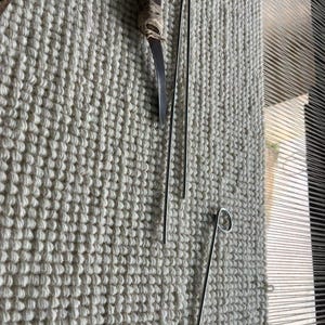 Puede incluir: Una herramienta con mango de madera marr&oacute;n oscuro y hoja de metal curva, dos varillas de metal plateado largas y delgadas, y una varilla de metal plateado con un bucle circular en un extremo, sobre una tela tejida de color blanco roto.