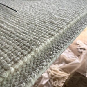 Puede incluir: Una alfombra de lana color blanco roto tejida a mano con una construcci&oacute;n densa de pelo rizado texturizado. El tejido muestra un patr&oacute;n consistente y repetitivo de peque&ntilde;os nudos circulares elevados en toda la superficie, creado en un telar.
