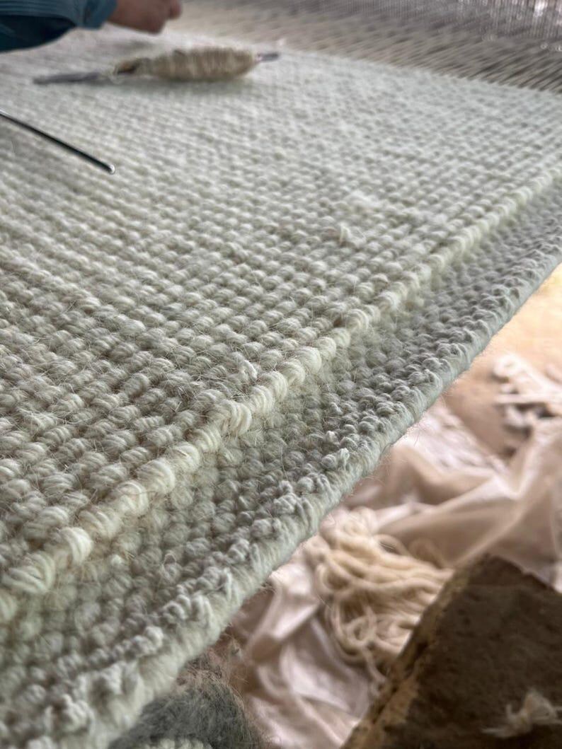 Puede incluir: Una alfombra de lana color blanco roto tejida a mano con una construcci&oacute;n densa de pelo rizado texturizado. El tejido muestra un patr&oacute;n consistente y repetitivo de peque&ntilde;os nudos circulares elevados en toda la superficie, creado en un telar.