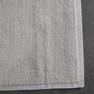 Puede incluir: Una alfombrilla de ba&ntilde;o rectangular de color blanco roto que presenta un patr&oacute;n de tejido de gofre texturizado en la superficie. La alfombrilla tiene un borde distintivo, liso y tejido plano que enmarca el dise&ntilde;o central de rejilla.