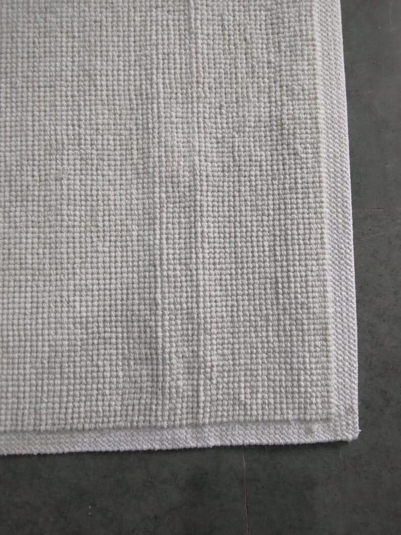 Puede incluir: Una alfombrilla de ba&ntilde;o rectangular de color blanco roto que presenta un patr&oacute;n de tejido de gofre texturizado en la superficie. La alfombrilla tiene un borde distintivo, liso y tejido plano que enmarca el dise&ntilde;o central de rejilla.