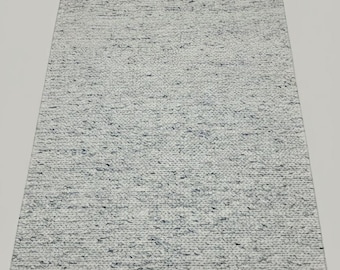 Puede incluir: Una alfombra rectangular gris claro con una superficie texturizada. La alfombra tiene un aspecto tejido con sutiles motas azul oscuro. La alfombra se muestra sobre un fondo blanco.