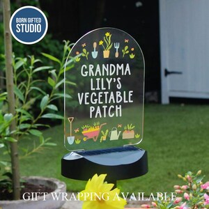 May include: A clear acrylic garden stake with the text "Grandma Lily's Vegetable Patch" and garden-themed illustrations. The stake is mounted on a black solar-powered base, ideal for outdoor use. The Born Gifted Studio logo is in the upper left corner.
