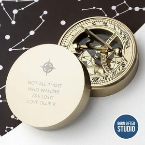 May include: A brass sundial compass with an open lid. The lid is engraved with a compass rose and the text "NOT ALL THOSE WHO WANDER ARE LOST! LOVE OLLIE X." The compass face has Roman numerals and a gnomon.