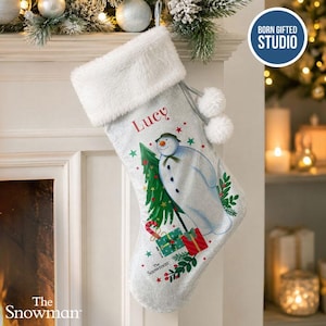 May include: A light grey Christmas stocking with a white faux fur cuff and two white pom-poms. The stocking features a printed illustration of a snowman holding a green Christmas tree, colourful gift boxes, and the name Lucy in red text.