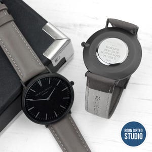 May include: Two black wristwatches with grey leather straps are shown. One watch face is visible, the other shows the engraved message: "WORLD'S BEST DAD! LOTS OF LOVE OLIVER X." The watches are on a white surface.