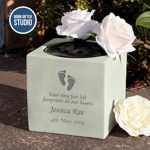 May include: A light green memorial vase with a dark top, holding white roses. The vase features engraved baby footprints and the text "Your tiny feet left footprints on our hearts Jessica Rae 4th May 2019". The "Born Gifted Studio" logo is in the top left.