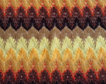 May include: Close-up of a knitted textile with a repeating chevron pattern. The color palette includes shades of yellow, orange, brown, and burgundy. The texture appears soft and the design is intricate, creating a visually appealing surface.