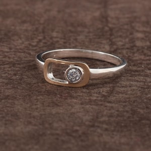 May include: A silver ring with a gold-coloured rectangular accent. The ring features a small, round, clear gemstone set in the centre of the gold accent. The ring is displayed on a brown surface.