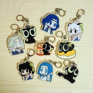 May include: A collection of cartoon character keychains with silver key rings. The keychains feature various anime-style characters, including cats and figures with blue, white, and black hair. Some keychains have text. The keychains are on a light wood-grain surface.