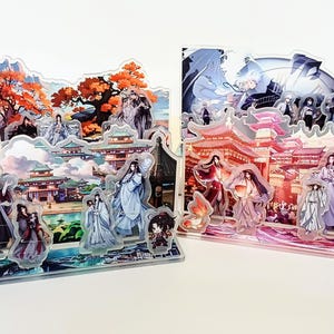 May include: Two acrylic display stands with anime-style illustrations. One shows a landscape with red and orange trees, the other a cityscape with figures in traditional dress. The stands are transparent with detailed artwork.