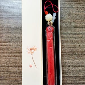 Può includere: Un ornamento a nappa rosso con una parte superiore decorativa, perline e un cordino rosso, presentato in una scatola color crema con un motivo floreale. La nappa è racchiusa in plastica trasparente.