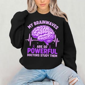Puede incluir: Sudadera negra con un gráfico de cerebro morado y el texto "MY BRAINWAVES ARE SO POWERFUL DOCTORS STUDY THEM". El diseño incluye una ilustración de cerebro, una línea de latidos y cintas moradas.