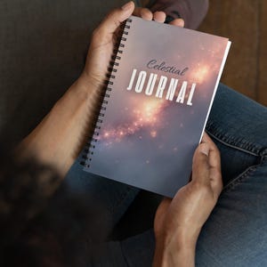 Puede incluir: Un diario con espiral sostenido en las manos de una persona. La portada presenta las palabras "Celestial JOURNAL" en blanco, con un diseño de galaxia en tonos morados, rosas y naranjas. El diario está abierto y la persona lleva vaqueros azules.