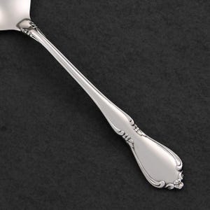 Op de afbeelding: Een zilveren serveerlepel met een decoratief bloemmotief op het handvat. De lepel heeft een gebogen, langwerpig handvat en een brede, platte kom. De lepel is gemaakt van glanzend, reflecterend metaal en staat tegen een donkere achtergrond.