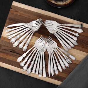 Puede incluir: Un surtido de tenedores plateados con diseños ornamentados en los mangos, dispuestos sobre una tabla de cortar de madera. Los tenedores tienen varios patrones, incluidos motivos florales y geométricos, y se muestran en forma de abanico.