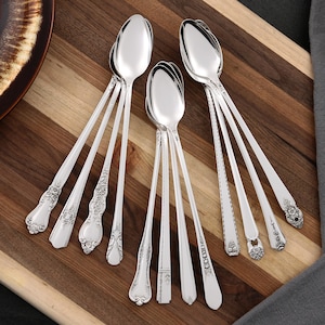 Puede incluir: Una colección de cucharas plateadas con diseños ornamentados en los mangos, dispuestas sobre una tabla de cortar de madera. Las cucharas presentan varios patrones, incluyendo motivos florales y geométricos. Las cucharas son brillantes y parecen ser de acero inoxidable.