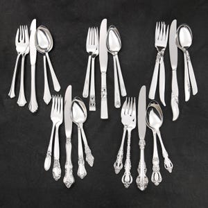 Op de afbeelding: Een assortiment zilverkleurig bestek, waaronder vorken, messen en lepels, gerangschikt op een donkere ondergrond. Het bestek heeft verschillende handvatontwerpen, van eenvoudig tot sierlijk, en toont een reeks eettijlstijlen.