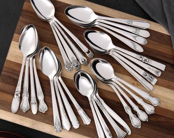 Può includere: Una collezione di cucchiai d'argento con disegni di manici ornati, disposti su un tagliere di legno. I cucchiai variano nello stile, con alcuni con motivi floreali e geometrici. I cucchiai sono riflettenti e la disposizione suggerisce un ambiente formale.