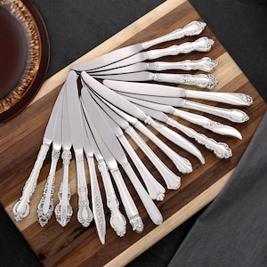 Puede incluir: Conjunto de cuchillos de mantequilla plateados con mangos ornamentados, dispuestos sobre una tabla de madera. Los cuchillos tienen diseños decorativos y hojas pulidas. La disposición sugiere una mesa formal.