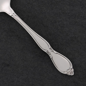May include: A close-up of a silver spoon with a floral design on the handle. The spoon has a smooth, polished finish and a decorative floral pattern at the base of the handle. The spoon is set against a dark background.
