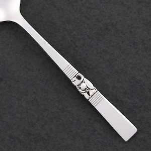 May include: Close-up of a silver spoon with a decorative floral design on the handle. The spoon has a rectangular end and a curved bowl. The handle features horizontal lines and a detailed floral pattern.