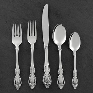 May include: A collection of five silver-coloured pieces of cutlery displayed on a dark grey surface. The set includes two forks, a knife, and two spoons. The handles have an ornate, floral design.