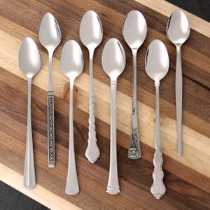 Puede incluir: Un surtido de ocho cucharas plateadas con varios dise&ntilde;os de mango, incluyendo patrones ornamentados y estilos simples y elegantes. Las cucharas est&aacute;n dispuestas sobre una tabla de cortar de madera, mostrando sus diferentes formas y tama&ntilde;os. Ideal para una cocina o comedor.