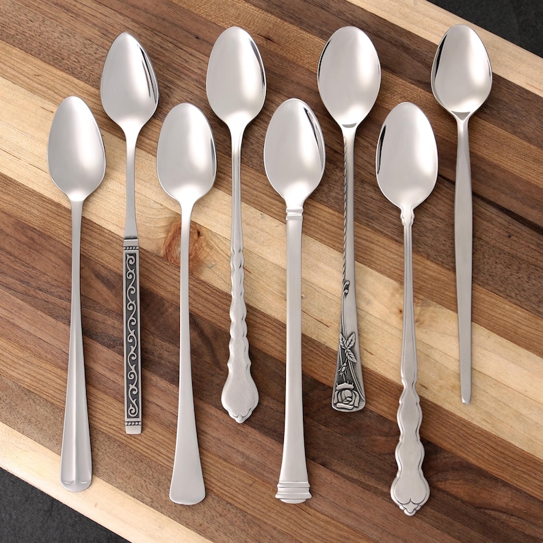 Puede incluir: Un surtido de ocho cucharas plateadas con varios dise&ntilde;os de mango, incluyendo patrones ornamentados y estilos simples y elegantes. Las cucharas est&aacute;n dispuestas sobre una tabla de cortar de madera, mostrando sus diferentes formas y tama&ntilde;os. Ideal para una cocina o comedor.