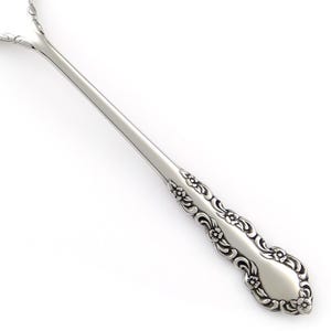 May include: A silver-toned metal spoon handle featuring an embossed floral and scrollwork pattern along the edges. The design includes small flower motifs integrated into the decorative border, extending from the base up to the neck of the spoon.