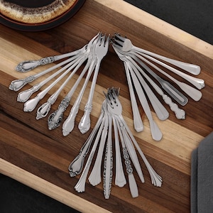 Puede incluir: Una variedad de tenedores plateados con diversos diseños de mango, incluyendo patrones florales ornamentados y estilos simples y elegantes. Los tenedores están dispuestos sobre una bandeja de madera, mostrando sus diferentes formas y texturas. La imagen destaca la variedad de cubiertos.