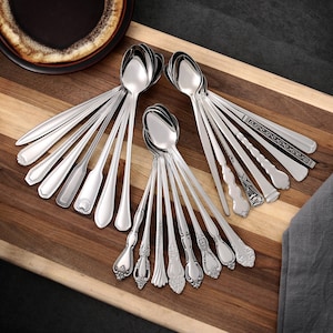 Puede incluir: Una colección de cucharas plateadas brillantes con varios diseños de mango, dispuestas sobre una tabla de cortar de madera. Las cucharas tienen diferentes patrones, incluyendo estilos florales, geométricos y clásicos simples. Un plato marrón oscuro es visible en la esquina superior izquierda.