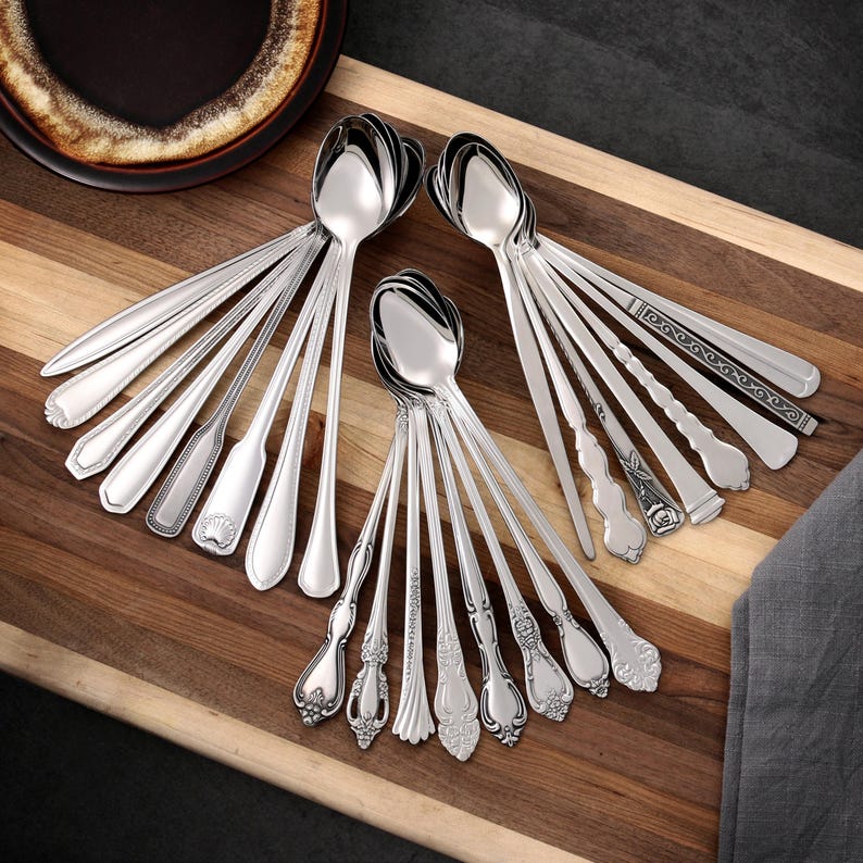 Puede incluir: Una colecci&oacute;n de cucharas plateadas brillantes con varios dise&ntilde;os de mango, dispuestas sobre una tabla de cortar de madera. Las cucharas tienen diferentes patrones, incluyendo estilos florales, geom&eacute;tricos y cl&aacute;sicos simples. Un plato marr&oacute;n oscuro es visible en la esquina superior izquierda.