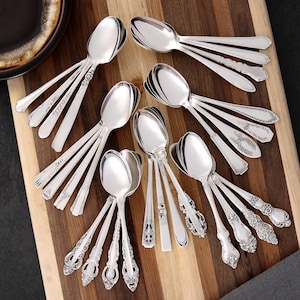 May include: An assortment of silver spoons with various handle designs, including ornate floral patterns and simple, elegant lines. The spoons are arranged on a wooden cutting board, showcasing their polished surfaces and reflective qualities.