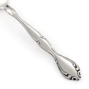 May include: A silver-toned metal spoon handle featuring a polished finish and a decorative embossed scroll pattern along the edges. The design includes a central open cutout section and a tapered end with symmetrical curved detailing.