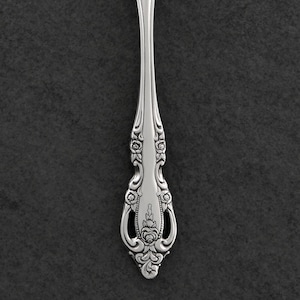 Könnte beinhalten: Nahaufnahme eines silbernen Löffels mit aufwendigen floralen Details. Der Löffelstiel weist ein symmetrisches Design aus Rosen und Scrollmustern auf. Das Besteck ist vor einem dunkelgrauen Hintergrund platziert, der die komplizierten Details hervorhebt.