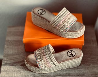 May include: A pair of platform sandals with woven straps in beige, pink, and blue. The sandals have a jute-style sole and a logo on the footbed. They are displayed on a wooden surface with an orange box.