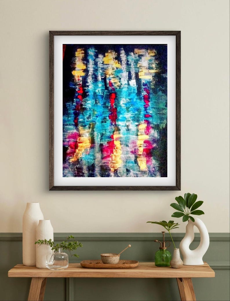 May include: Abstract art print in a dark wooden frame. The artwork features vertical brushstrokes in shades of blue, red, yellow, and pink against a dark background. The painting is displayed above a wooden table with decorative vases.