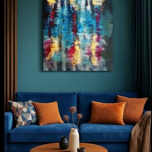 May include: An abstract painting with blue, yellow, and red brushstrokes hangs above a blue velvet sofa. The sofa is adorned with orange throw pillows and a patterned pillow. A round wooden coffee table holds a vase, small black vases, and a bowl.
