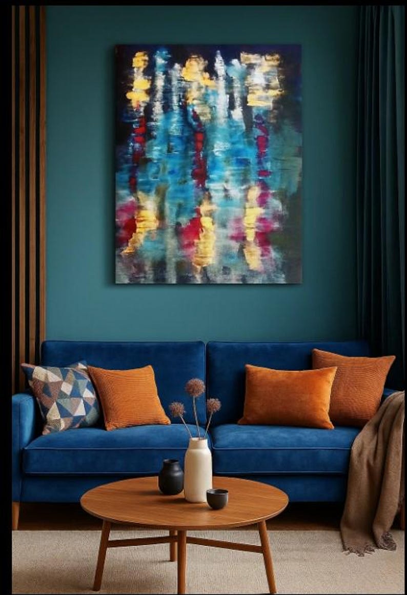 May include: An abstract painting with blue, yellow, and red brushstrokes hangs above a blue velvet sofa. The sofa is adorned with orange throw pillows and a patterned pillow. A round wooden coffee table holds a vase, small black vases, and a bowl.