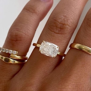 May include: A hand displays four gold rings. One ring features a rectangular, clear gemstone. Another ring has a curved design with small, clear stones. The remaining rings are simple bands. The rings are set against a neutral background.