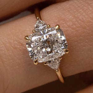 Peut inclure: Une bague en or avec un gros diamant central de taille carrée, flanqué de deux diamants triangulaires plus petits. Le bracelet de la bague est en or poli. Les diamants scintillent. Un bijou.