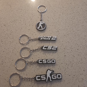 Puede incluir: Llaveros con el logotipo y los personajes del videojuego Counter-Strike. Los llaveros tienen una combinación de colores blanco y negro. Un llavero tiene un diseño circular con una figura silueteada sosteniendo un arma. Otros llaveros tienen el texto "Counter Strike 2", "CS2" y "CSGO".