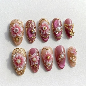 May include: A set of ten artificial nails with a variety of designs. The nails feature pink and gold floral embellishments, pearls, and glitter. Some nails have a solid pink base, while others have a nude base.
