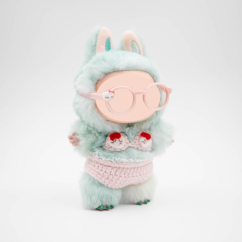 Puede incluir: Una figura de conejito de peluche con un conjunto con capucha de piel verde menta, gafas rosas y un bikini rosa de ganchillo. La parte superior del bikini tiene detalles en rojo. La figura tiene una cara en blanco y pies verdes.