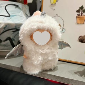 Pode incluir: Um brinquedo de pelúcia branco e fofo em forma de criatura, com pequenos membros e asas prateadas. Possui um rosto em forma de coração e um pequeno adorno de cabeça rosa e prateado.