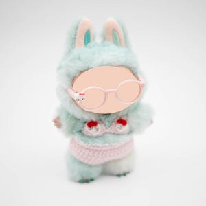 Puede incluir: Mu&ntilde;eca de peluche de conejito con un atuendo con capucha azul claro con detalles en rosa y un bikini de ganchillo rosa. La mu&ntilde;eca tiene gafas rosas redondas y una cara en blanco. Las orejas de conejo est&aacute;n forradas con tela turquesa.