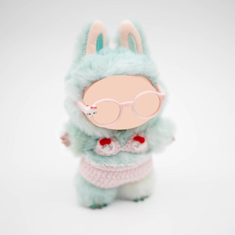 Puede incluir: Mu&ntilde;eca de peluche de conejito con un atuendo con capucha azul claro con detalles en rosa y un bikini de ganchillo rosa. La mu&ntilde;eca tiene gafas rosas redondas y una cara en blanco. Las orejas de conejo est&aacute;n forradas con tela turquesa.