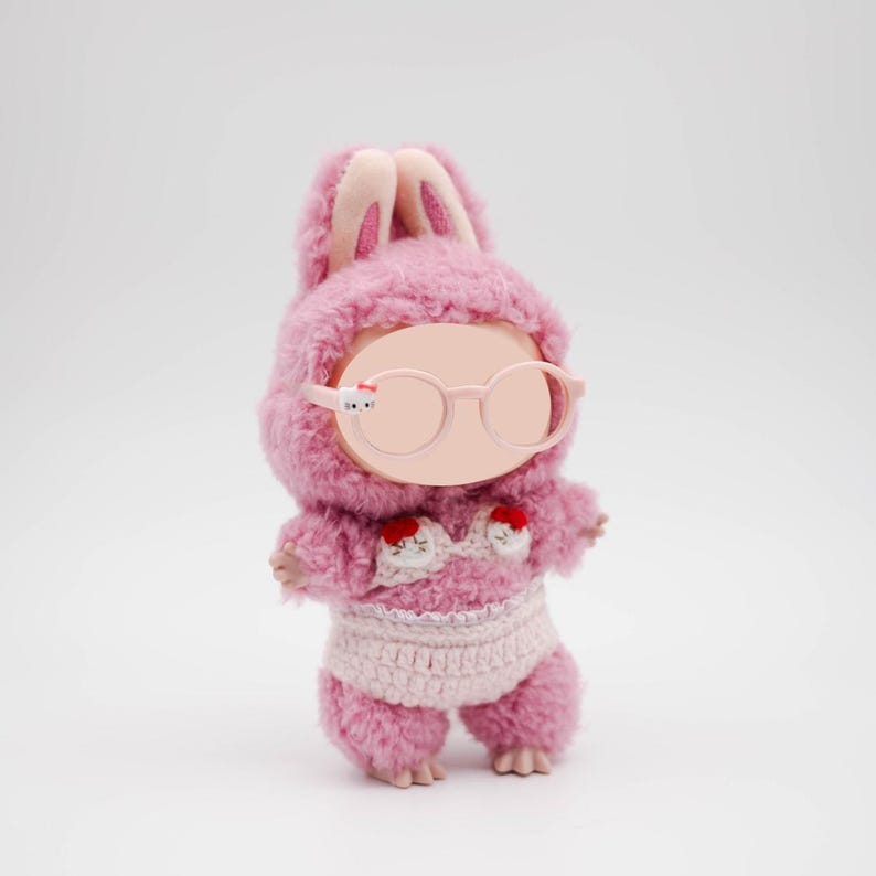 Puede incluir: Mu&ntilde;eca de peluche rosa con un conjunto con capucha y gafas a juego, y un bikini de ganchillo. La mu&ntilde;eca tiene orejas rosas y pies peque&ntilde;os. El bikini tiene detalles rojos. La mu&ntilde;eca est&aacute; sobre un fondo blanco.