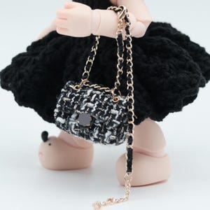 May include: A miniature black and white tweed handbag with a gold chain strap. The small purse has a black flower-shaped clasp and is held by a doll. The doll is wearing a black dress.